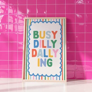 May include: A framed art print with the words "BUSY DILLY DALLY ING" in colorful block letters. The print has a white background with a scalloped border and a rainbow striped frame. The background is a bright pink tiled wall.
