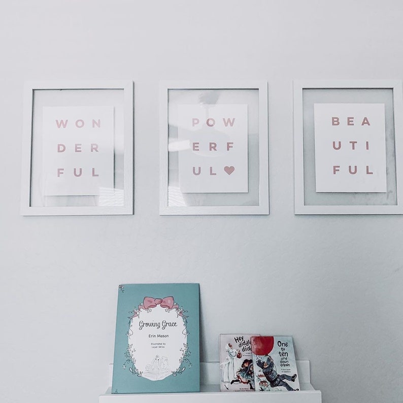 May include: Three white picture frames with pink lettering spelling out "Wonderful", "Powerful", and "Beautiful". The frames are hanging on a white wall. Below the frames are three books: "Growing Grace" by Erin Mason, "Hey diddl diddl" and "One to ten again".