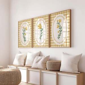 May include: Three framed prints featuring yellow flowers on a white background. Each print has a gingham pattern in yellow and white. The prints are hanging on a white wall above a wooden bench with white cushions.