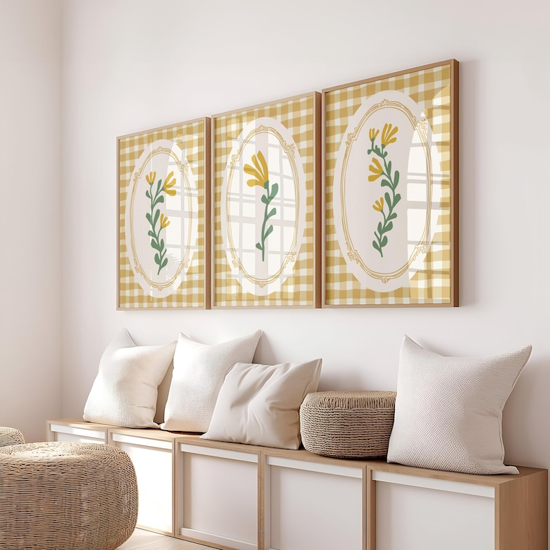 May include: Three framed prints featuring yellow flowers on a white background. Each print has a gingham pattern in yellow and white. The prints are hanging on a white wall above a wooden bench with white cushions.