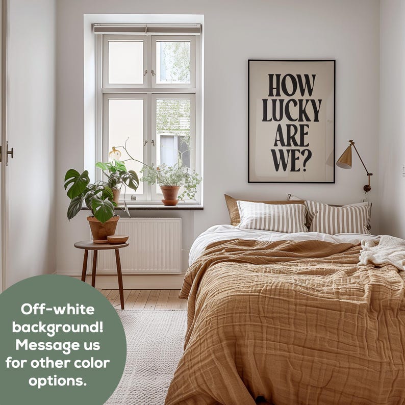 May include: A bedroom with a white wall and a window. A bed with a brown blanket and white and brown striped pillows is in the center of the room. A black framed poster with the text "HOW LUCKY ARE WE?" is hanging on the wall above the bed. A small table with a potted plant is next to the window.