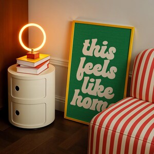 May include: A cream-colored bedside table with two drawers, topped with books and a circular lamp. A framed green poster with the words "this feels like home" leans against the wall. A striped armchair is in the corner.
