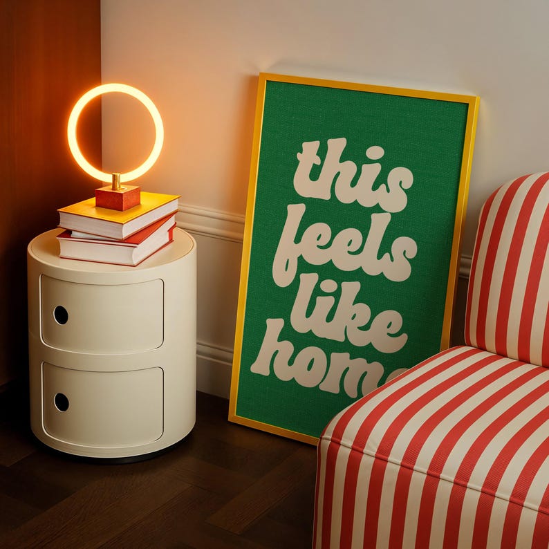 May include: A cream-colored bedside table with two drawers, topped with books and a circular lamp. A framed green poster with the words "this feels like home" leans against the wall. A striped armchair is in the corner.