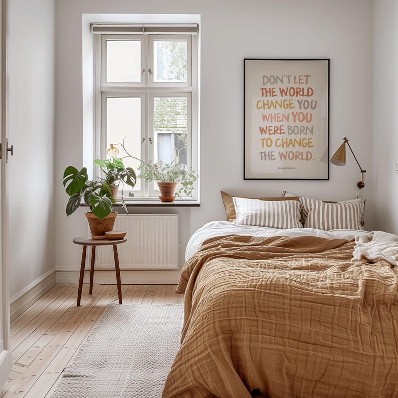 K&ouml;nnte beinhalten: Ein Schlafzimmer mit einem Bett mit einer braunen Decke und wei&szlig;en und grauen gestreiften Kissen. Ein gerahmtes Bild mit dem Text "Lass die Welt dich nicht ver&auml;ndern, wenn du geboren wurdest, um die Welt zu ver&auml;ndern." h&auml;ngt an der Wand &uuml;ber dem Bett.