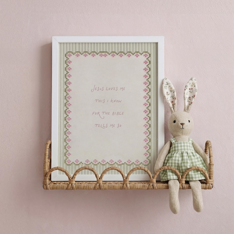 May include: A white framed cross-stitch art piece featuring a pink floral border and the text Jesus loves me this I know for the bible tells me so, displayed on a woven rattan shelf next to a plush rabbit toy wearing a green gingham dress.