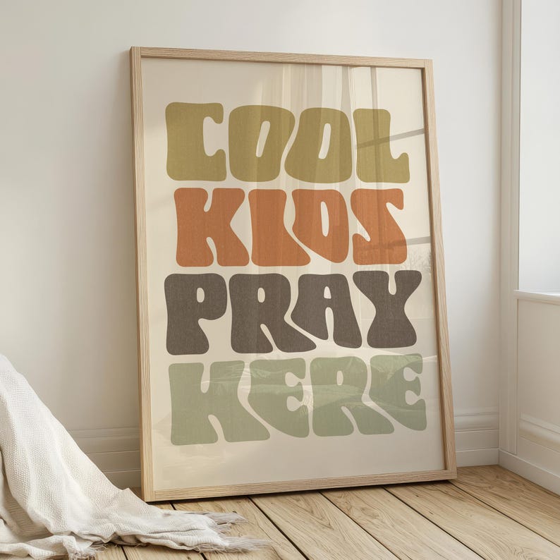 May include: A framed art print with the words "COOL KIDS PRAY HERE" in large, retro-style lettering. The text is in shades of olive green, orange, and brown against a cream background. The frame is a light wood color.