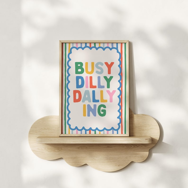 May include: A framed art print with the words "BUSY DILLY DALLY ING" in colorful letters. The print has a white background with a scalloped border and is displayed on a cloud-shaped wooden shelf.