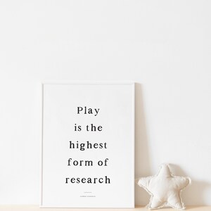 May include: A white framed print with the quote "Play is the highest form of research" by Albert Einstein. A white star-shaped pillow sits on a light beige surface.