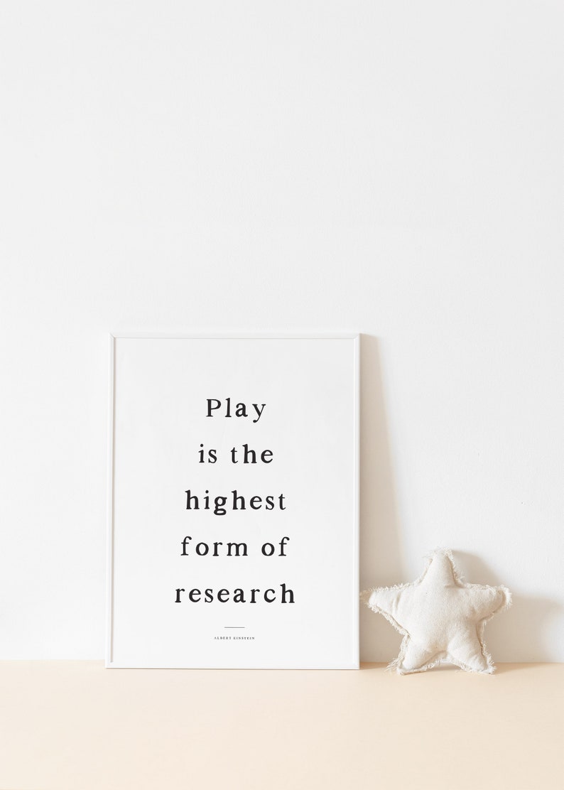 May include: A white framed print with the quote "Play is the highest form of research" by Albert Einstein. A white star-shaped pillow sits on a light beige surface.