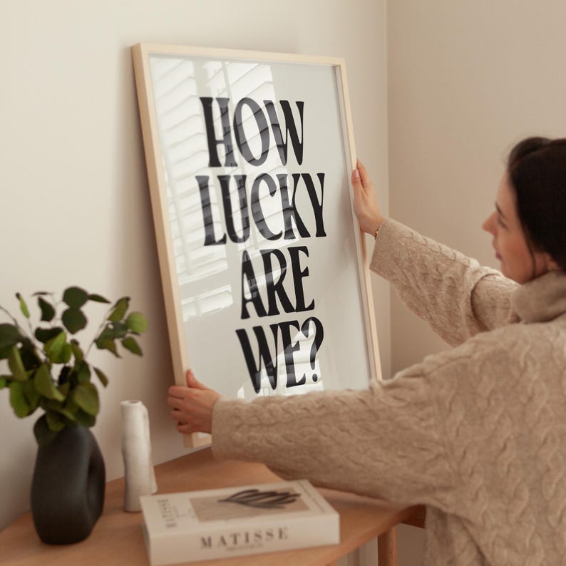 May include: A framed print with the text "How lucky are we?" in black bold letters on a white background. The print is being hung on a wall by a person.