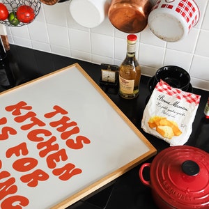 May include: A framed print with the text "This Kitchen Is For Zing" in orange letters on a white background. The print is hanging on a black countertop with a red Le Creuset Dutch oven, a bag of pastries, a bottle of wine, and a variety of kitchen utensils.