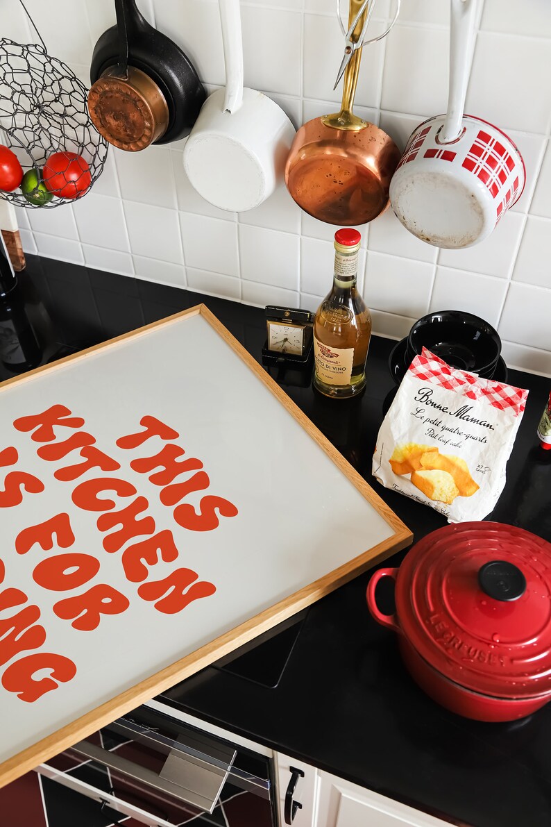 May include: A framed print with the text "This Kitchen Is For Zing" in orange letters on a white background. The print is hanging on a black countertop with a red Le Creuset Dutch oven, a bag of pastries, a bottle of wine, and a variety of kitchen utensils.