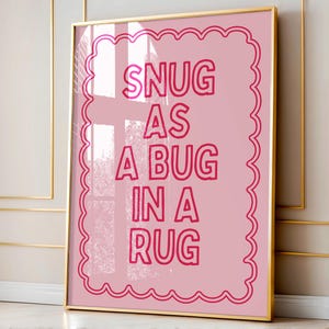 Peut inclure: Impression artistique encadrée, rose, avec l'inscription "SNUG AS A BUG IN A RUG" en rouge. Le texte est centré sur un fond rose avec une bordure festonnée. Le cadre est doré.