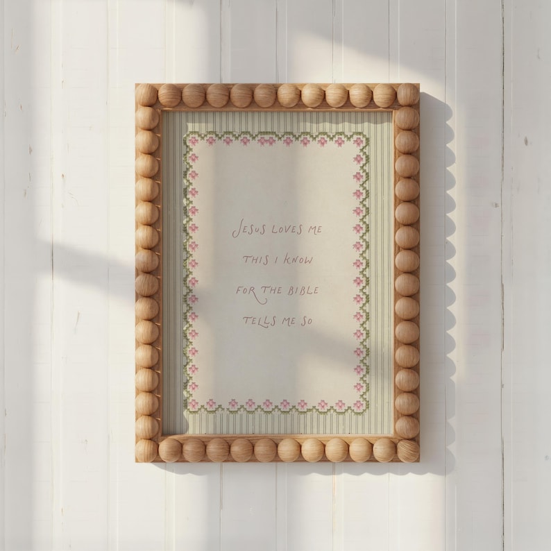 May include: A rectangular wall art piece in a light wood frame with a beaded border. The print features a cream-colored center with a pink and green floral cross-stitch border and the text: Jesus loves me, this I know, for the Bible tells me so.