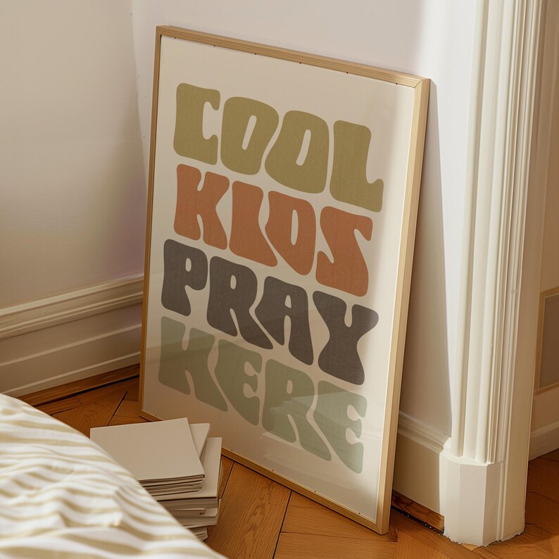 May include: A framed art print with the words "COOL KIDS PRAY HERE" in a retro font. The text is in shades of green, orange, and brown on a cream background. The frame is a light wood color. The print is leaning against a wall.