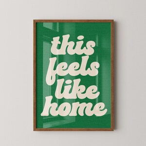 May include: A framed art print with the words "this feels like home" in a cream-colored, retro font against a green background. The frame is a warm brown color, and the print is displayed on a white wall.