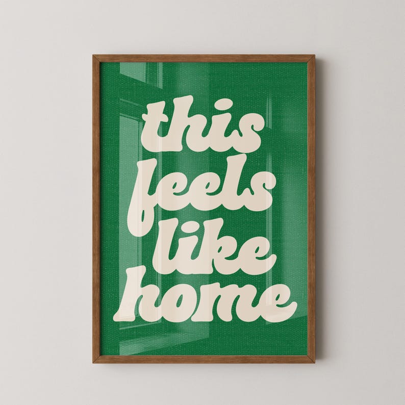 May include: A framed art print with the words "this feels like home" in a cream-colored, retro font against a green background. The frame is a warm brown color, and the print is displayed on a white wall.