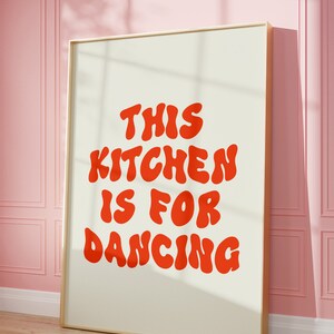 May include: A framed print with the text "This kitchen is for dancing" in red, bubble-like letters on a cream background.