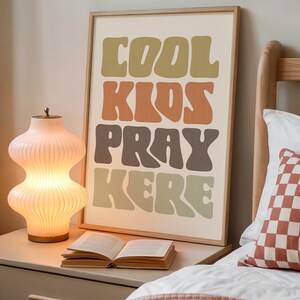 May include: A framed art print with the words "COOL KIDS PRAY HERE" in a retro font. The print is in a bedroom setting, next to a lit lamp and an open book. The color palette includes shades of green, orange, and brown.