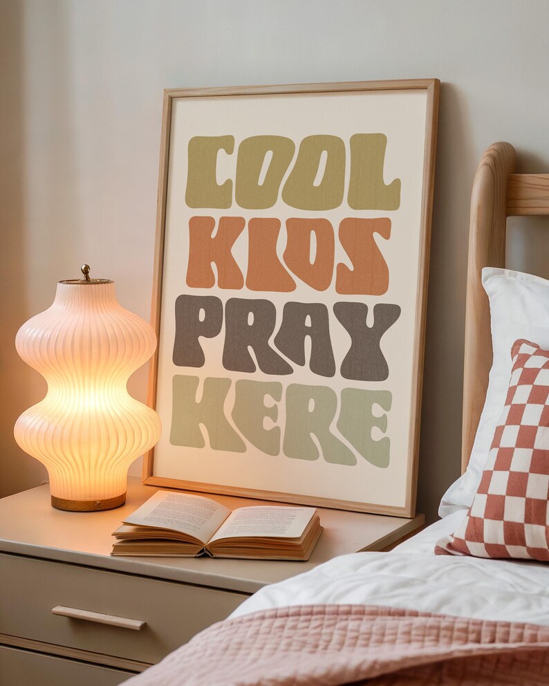 May include: A framed art print with the words "COOL KIDS PRAY HERE" in a retro font. The print is in a bedroom setting, next to a lit lamp and an open book. The color palette includes shades of green, orange, and brown.
