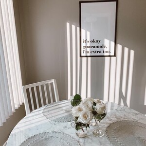 Puede incluir: Una mesa de m&aacute;rmol blanco con una impresi&oacute;n enmarcada en blanco que dice "it's okay guacamole, i'm extra too." La mesa est&aacute; puesta con manteles individuales blancos, un jarr&oacute;n blanco con flores blancas y dos copas de vidrio transparentes.