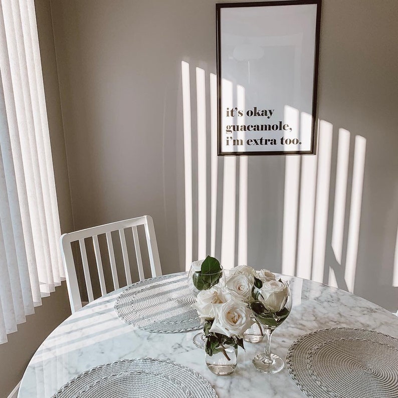 Puede incluir: Una mesa de m&aacute;rmol blanco con una impresi&oacute;n enmarcada en blanco que dice "it's okay guacamole, i'm extra too." La mesa est&aacute; puesta con manteles individuales blancos, un jarr&oacute;n blanco con flores blancas y dos copas de vidrio transparentes.
