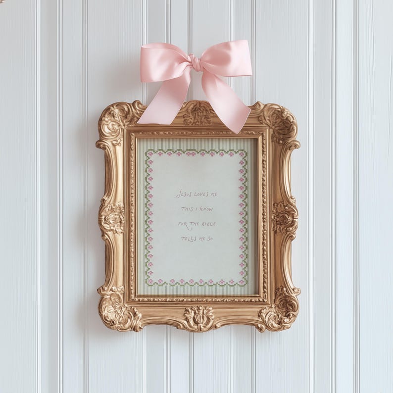May include: A rectangular gold ornate frame featuring a light pink satin bow at the top. Inside the frame is a light green background with a pink and green floral border and the text: Jesus loves me this I know for the Bible tells me so.