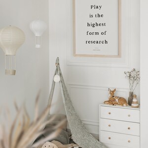 May include: A white dresser with three drawers and wooden knobs. Two small deer figurines are on top of the dresser, along with a wooden stacking toy. A framed print with the quote "Play is the highest form of research" hangs on the wall behind the dresser.