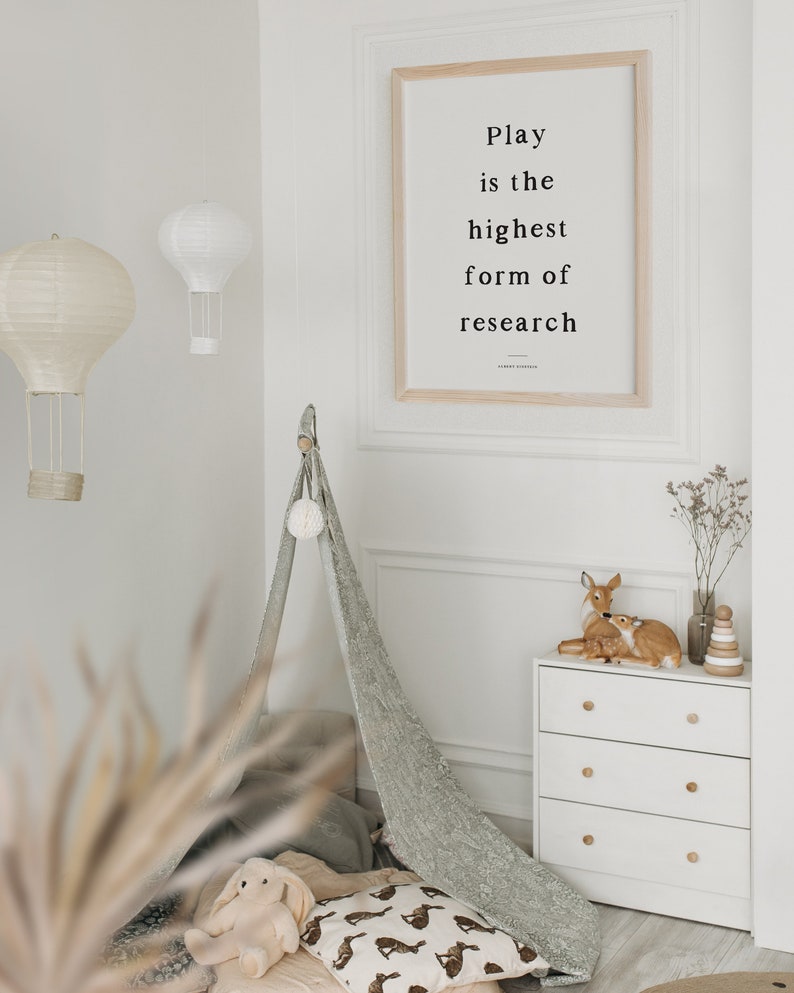 May include: A white dresser with three drawers and wooden knobs. Two small deer figurines are on top of the dresser, along with a wooden stacking toy. A framed print with the quote "Play is the highest form of research" hangs on the wall behind the dresser.