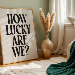 May include: A framed print with the text "HOW LUCKY ARE WE?" in black bold letters on a white background. The print is hanging on a light green wall in a room with a white rug, a vase with dried pampas grass, and a green throw blanket.