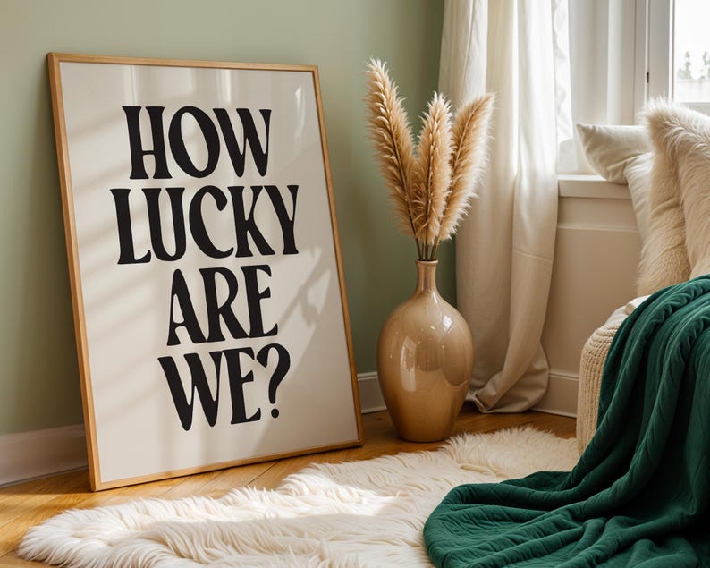 May include: A framed print with the text "HOW LUCKY ARE WE?" in black bold letters on a white background. The print is hanging on a light green wall in a room with a white rug, a vase with dried pampas grass, and a green throw blanket.