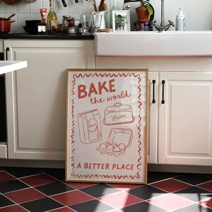 May include: A framed poster with a white background and red text that reads "BAKE the world a BETTER PLACE". The poster features a cartoon illustration of a bag of flour, a container of butter, and a carton of eggs.