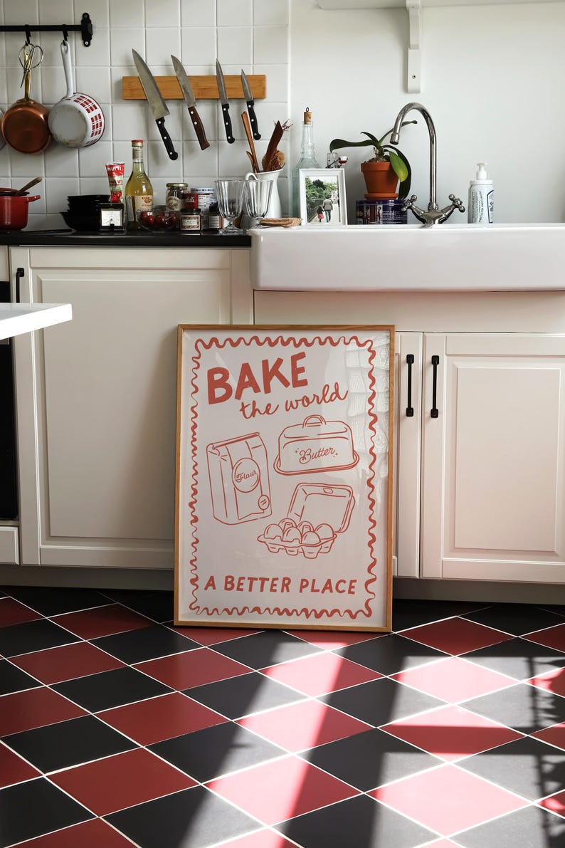 May include: A framed poster with a white background and red text that reads "BAKE the world a BETTER PLACE". The poster features a cartoon illustration of a bag of flour, a container of butter, and a carton of eggs.