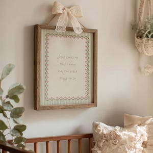 May include: A rectangular wooden framed wall hanging featuring a cream-colored fabric panel with a green striped border and a pink floral cross-stitch edge. The text reads Jesus loves me this I know for the bible tells me so. A lace bow is attached.