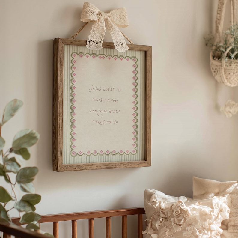 May include: A rectangular wooden framed wall hanging featuring a cream-colored fabric panel with a green striped border and a pink floral cross-stitch edge. The text reads Jesus loves me this I know for the bible tells me so. A lace bow is attached.