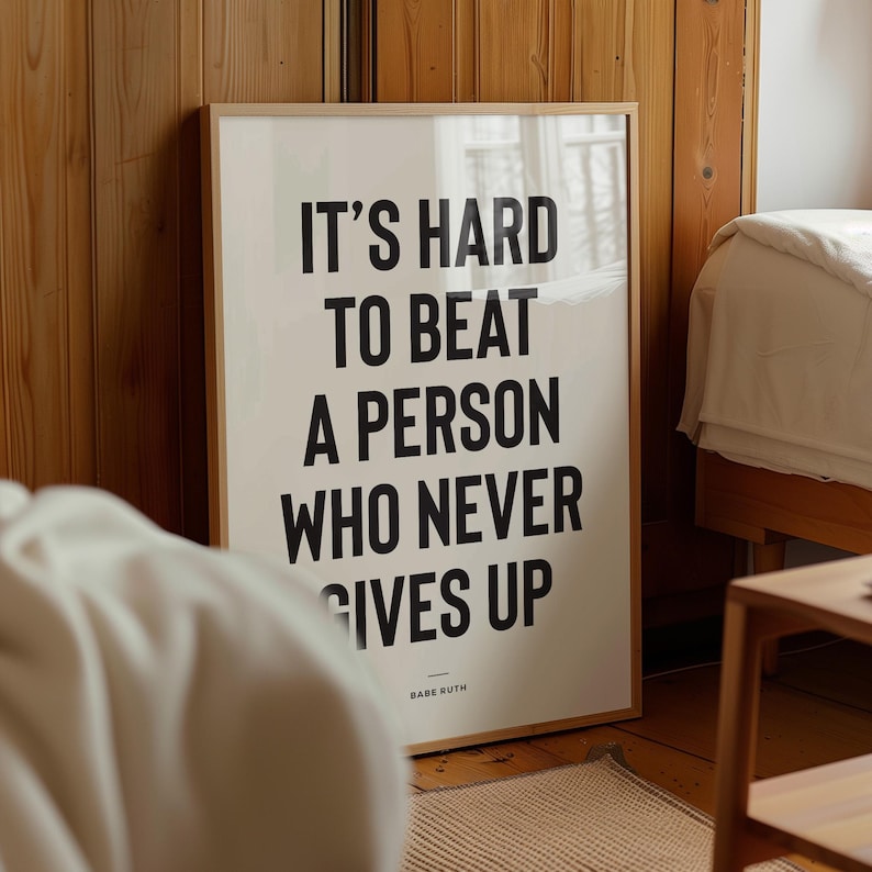 Puede incluir: Un p&oacute;ster enmarcado con la cita "IT'S HARD TO BEAT A PERSON WHO NEVER GIVES UP" en letras negras sobre un fondo crema. La cita se atribuye a Babe Ruth. El marco es de color madera clara.