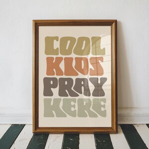 May include: A framed print with the words "COOL KIDS PRAY HERE" in a retro font. The text is in shades of green, orange, and brown against a cream background. The frame is a warm brown color.