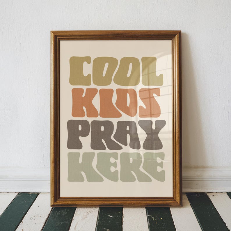 May include: A framed print with the words "COOL KIDS PRAY HERE" in a retro font. The text is in shades of green, orange, and brown against a cream background. The frame is a warm brown color.