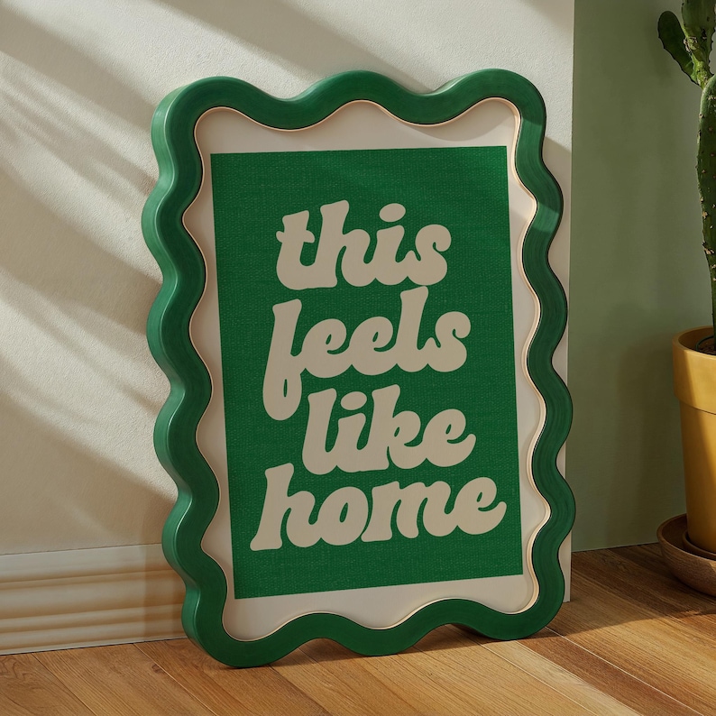May include: A framed wall art piece with a green wavy border and a white interior border. The artwork features the phrase "this feels like home" in a white, retro font against a green background. The frame is leaning against a wall.