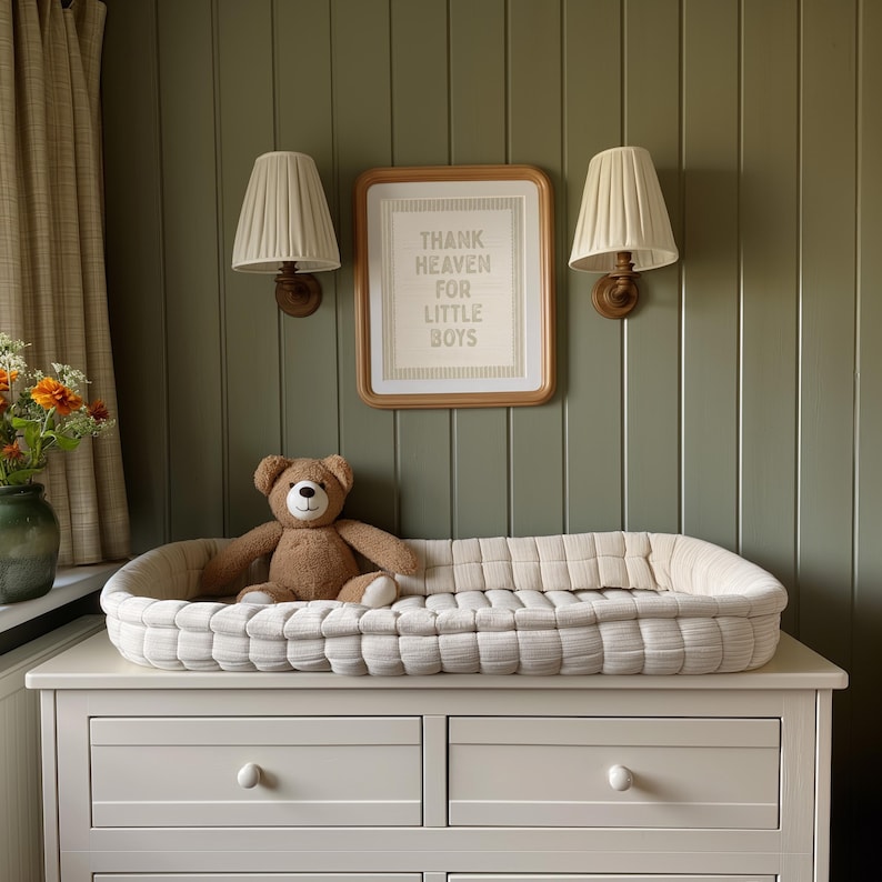 Puede incluir: Una escena de guarder&iacute;a con un cambiador blanco sobre una c&oacute;moda blanca y un oso de peluche marr&oacute;n. La pared verde oliva tiene un cartel enmarcado que dice "Thank Heaven for Little Boys". Dos l&aacute;mparas con pantallas crema est&aacute;n montadas.