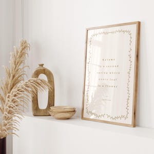 May include: A framed print with the quote "Autumn is a second spring where every leaf is a flower" in a light brown font. The frame is light brown, and the print is decorated with a floral border. Decorative wooden bowls and a vase are also visible.
