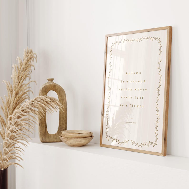 May include: A framed print with the quote "Autumn is a second spring where every leaf is a flower" in a light brown font. The frame is light brown, and the print is decorated with a floral border. Decorative wooden bowls and a vase are also visible.