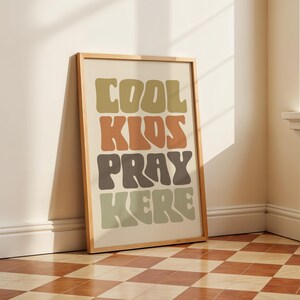 May include: A framed art print with the words "COOL KIDS PRAY HERE" in a retro font. The text is in shades of green, brown, and orange against a cream background. The wooden frame leans against a wall.
