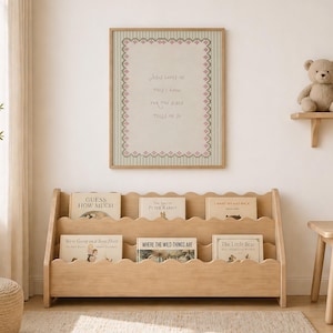May include: A light wood bookshelf with three scalloped tiers holding children's books. Above it hangs a framed print with a light green striped background, a pink decorative border, and the text: Jesus loves me this I know for the Bible tells me so.