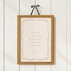 May include: A framed print with a gold-colored ornate frame and a green ribbon for hanging. The print features the quote "Autumn is a second spring where every leaf is a flower" in gold lettering, surrounded by a delicate floral border.