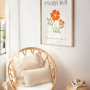 K&ouml;nnte beinhalten: Ein gerahmter Kunstdruck mit dem Text "PSALM 16:11" und orangefarbenen Blumenillustrationen. Der Druck h&auml;ngt &uuml;ber einem Korbstuhl mit cremefarbenen Kissen. Ein kleiner gewebter Hocker mit dekorativen Perlen und einer Quaste befindet sich neben dem Stuhl.