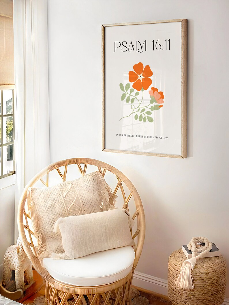 K&ouml;nnte beinhalten: Ein gerahmter Kunstdruck mit dem Text "PSALM 16:11" und orangefarbenen Blumenillustrationen. Der Druck h&auml;ngt &uuml;ber einem Korbstuhl mit cremefarbenen Kissen. Ein kleiner gewebter Hocker mit dekorativen Perlen und einer Quaste befindet sich neben dem Stuhl.