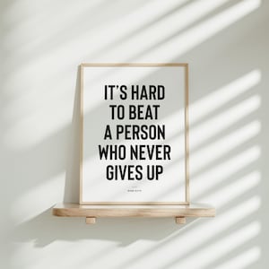 Puede incluir: Impresi&oacute;n enmarcada con la cita "IT'S HARD TO BEAT A PERSON WHO NEVER GIVES UP" en texto negro sobre fondo blanco. La impresi&oacute;n est&aacute; en un marco de madera clara y descansa sobre una estanter&iacute;a de madera.