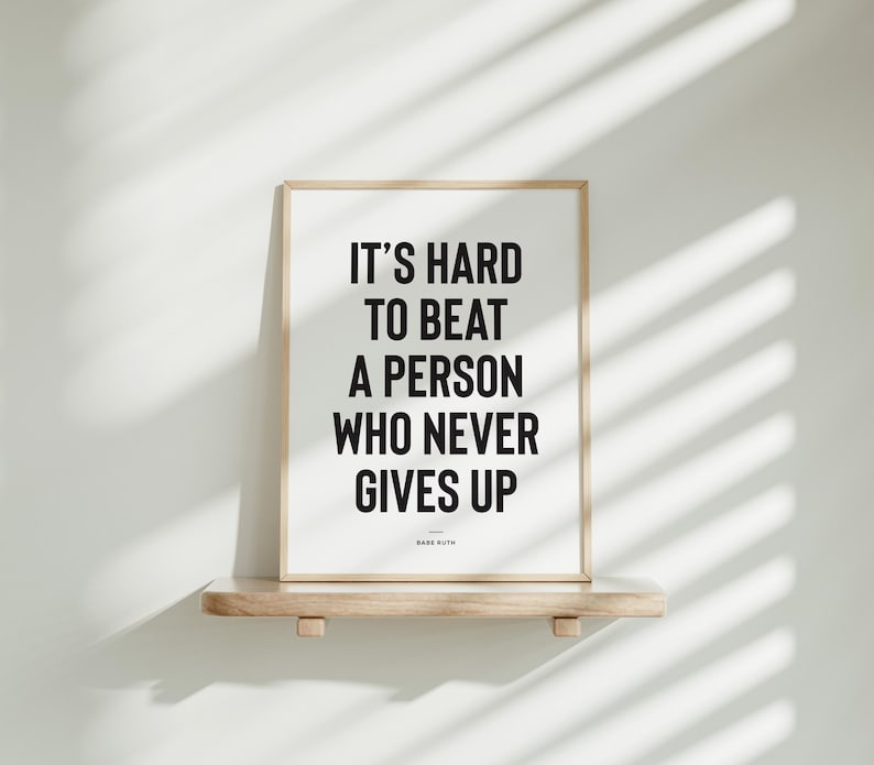 Puede incluir: Impresi&oacute;n enmarcada con la cita "IT'S HARD TO BEAT A PERSON WHO NEVER GIVES UP" en texto negro sobre fondo blanco. La impresi&oacute;n est&aacute; en un marco de madera clara y descansa sobre una estanter&iacute;a de madera.