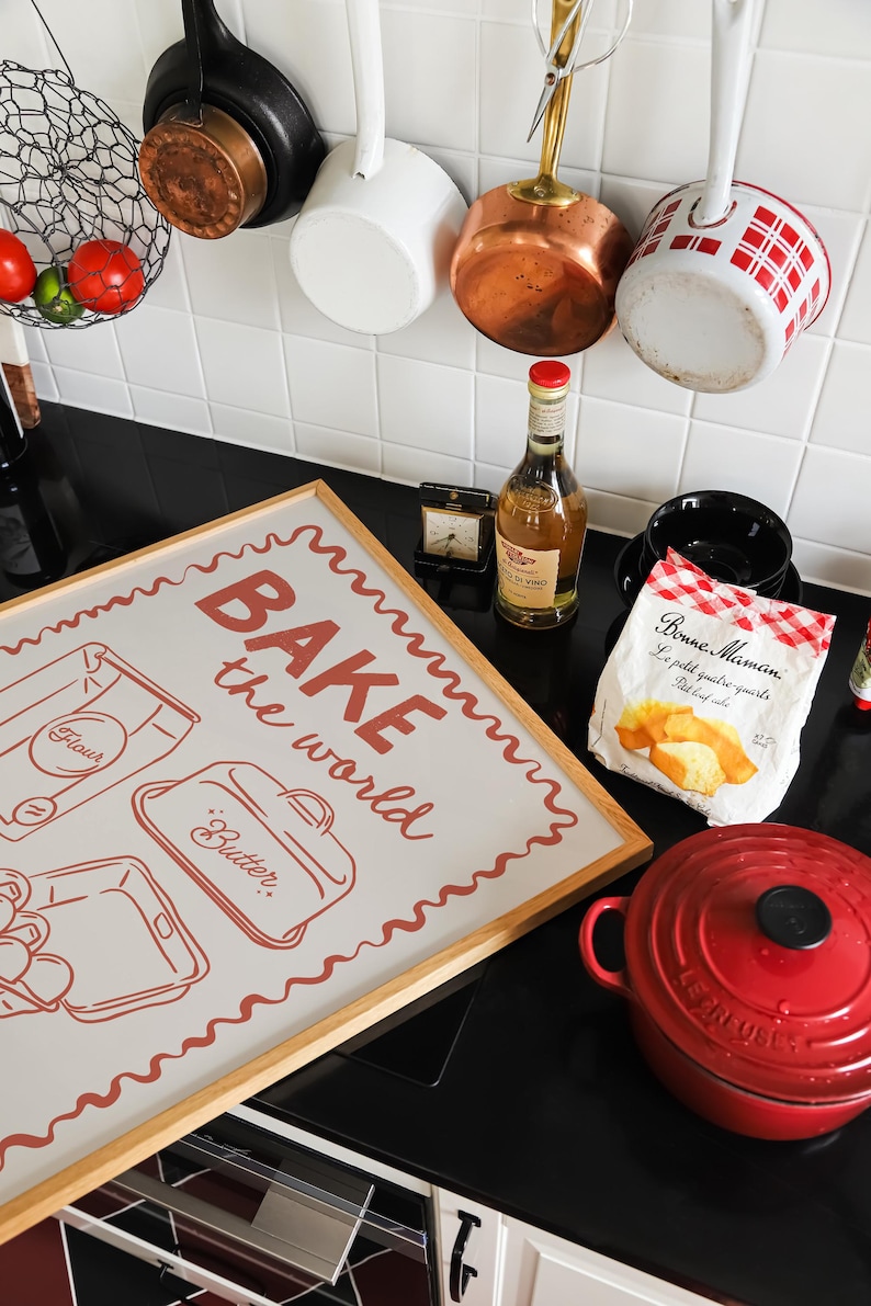 May include: A framed print with the text "BAKE the world" in red lettering on a light gray background. The print features illustrations of a bag of flour, a container of butter, and a baking pan. The print is hanging on a white tiled wall in a kitchen.